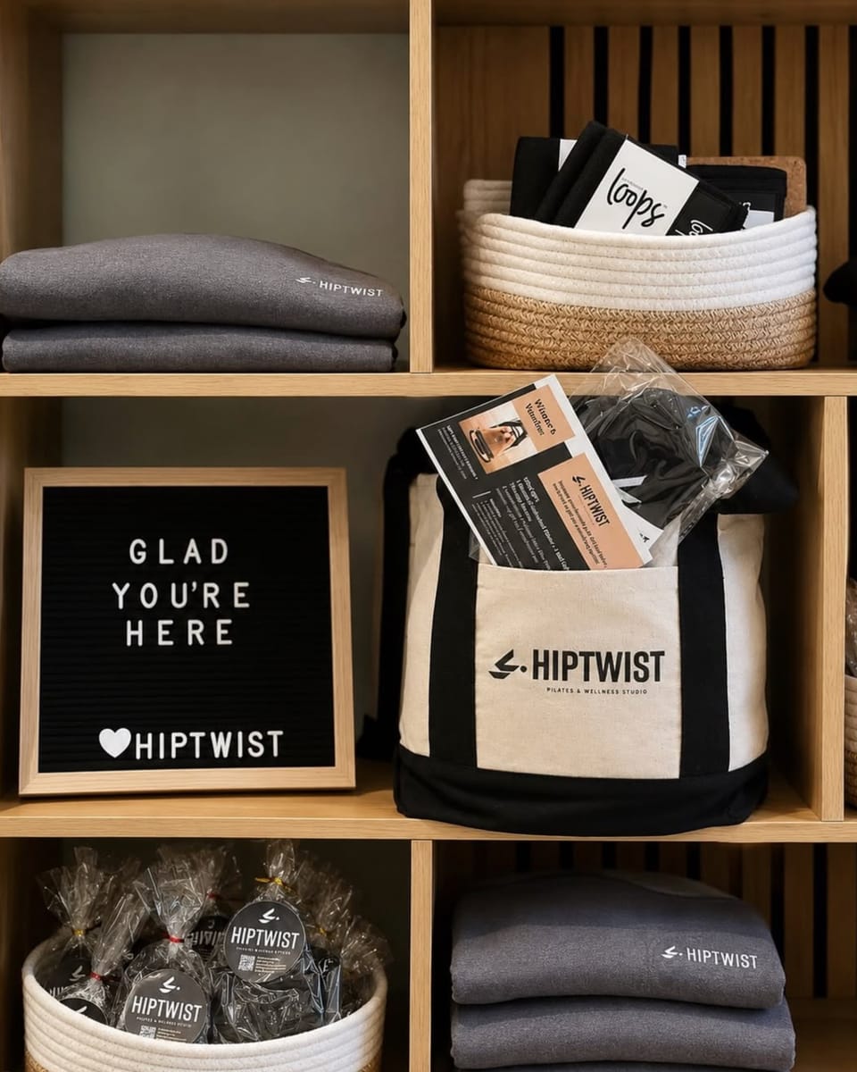 Shelves in the Hiptwist studio with folded sweatshirts, a Hiptwist tote bag, and a letterboard reading &ldquo;Glad you&rsquo;re here&rdquo;